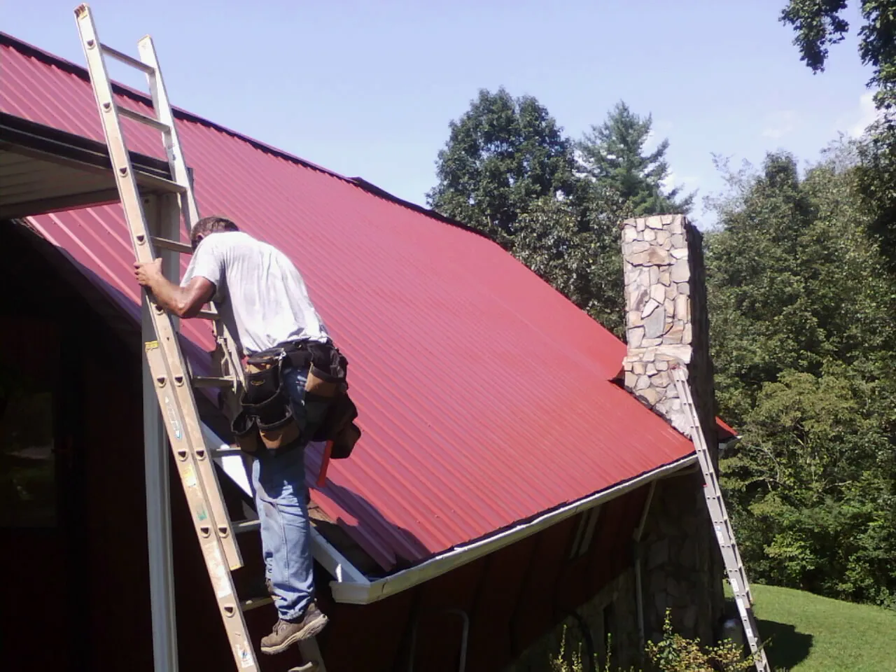 Roofer on a ladder beside a red metal roof and stone chimney — Soffit & Fascia Repair by Roofing Brothers in Baltimore